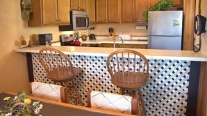 Een keuken met houten kastjes, een magnetron, een koelkast en twee rieten stoelen met kussens.