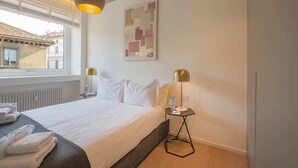 Ein Hotelzimmer mit einem Bett, zwei Nachttischlampen, einem Fenster mit Jalousien, einem Heizkörper und einem an der Wand hängenden abstrakten Gemälde.
