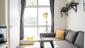 A modern living room with a sofa, a coffee table with a plant, a floor lamp, and a shelf with books and a plant.