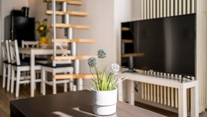 A modern living room with a wooden dining table, white chairs, a white vase with green plants, and a large flat-screen TV.