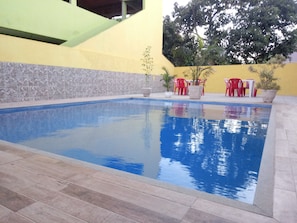A swimming pool with tiled flooring, surrounded by a yellow wall and a stone wall. There are red chairs and a table near the pool. - GUEST HOUSE RENASCER K&W (Cabo Frio)