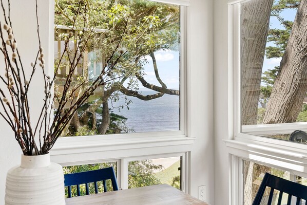 A room with a large window overlooking a body of water and trees, a white vase with branches, and a wooden table with blue chairs. - Bright, Steps to Sand, Ocean Views (La Selva Beach)