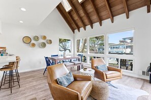 A spacious living area with a high ceiling, wooden beams, and large windows offering a view of the outside. - Bright, Steps to Sand, Ocean Views (La Selva Beach)