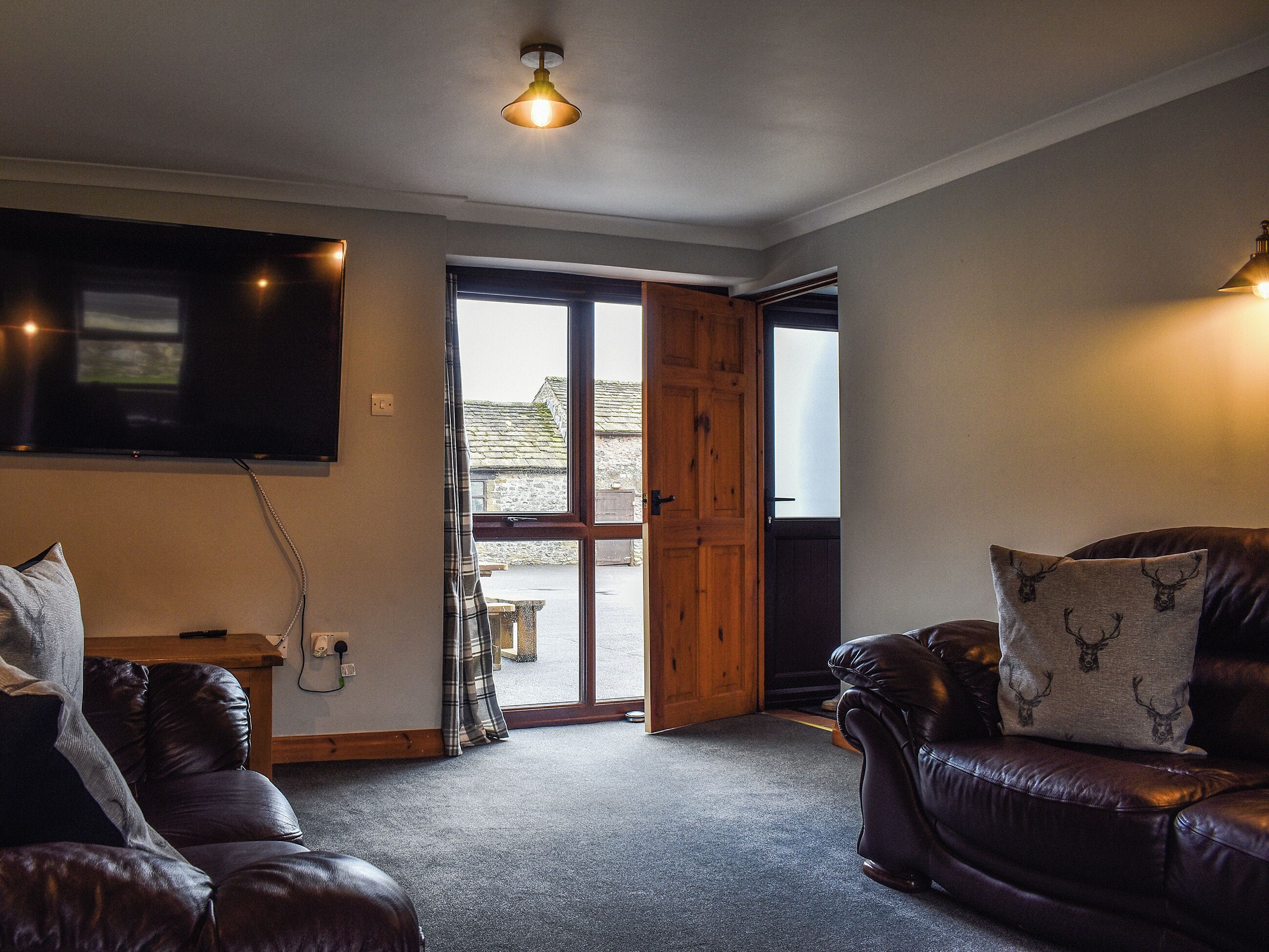 A cozy living room with a leather sofa, a wooden door leading outside, and a flat-screen TV mounted on the wall.