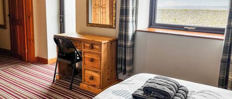 A hotel room with a bed, a desk, a chair, a wooden dresser, a mirror, and a window with a view of a vineyard.