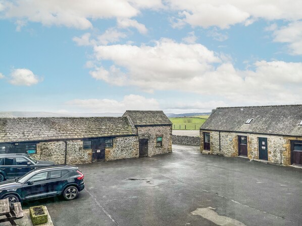 A stone building with a paved courtyard, a parked car, and a view of the countryside.