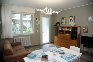 Ein Wohnzimmer mit einer Couch, einem Esstisch mit Platzsets, einer Bücherwand, einem Fernseher und einem Fenster mit Vorhängen.