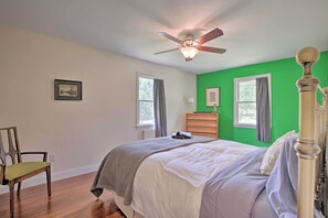 Un dormitorio con cama, una silla, cómoda, ventilador de techo y dos ventanas con cortinas.