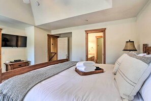 Een hotelkamer met een groot bed, een aan de muur bevestigde televisie, een houten nachtkastje en een deur naar een andere kamer.