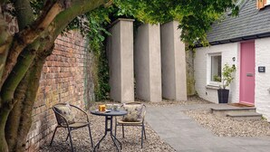 Ein Innenhof mit Backsteinmauer, einer Betonstruktur, einer rosafarbenen Tür und zwei Korbstühlen mit einem Tisch.