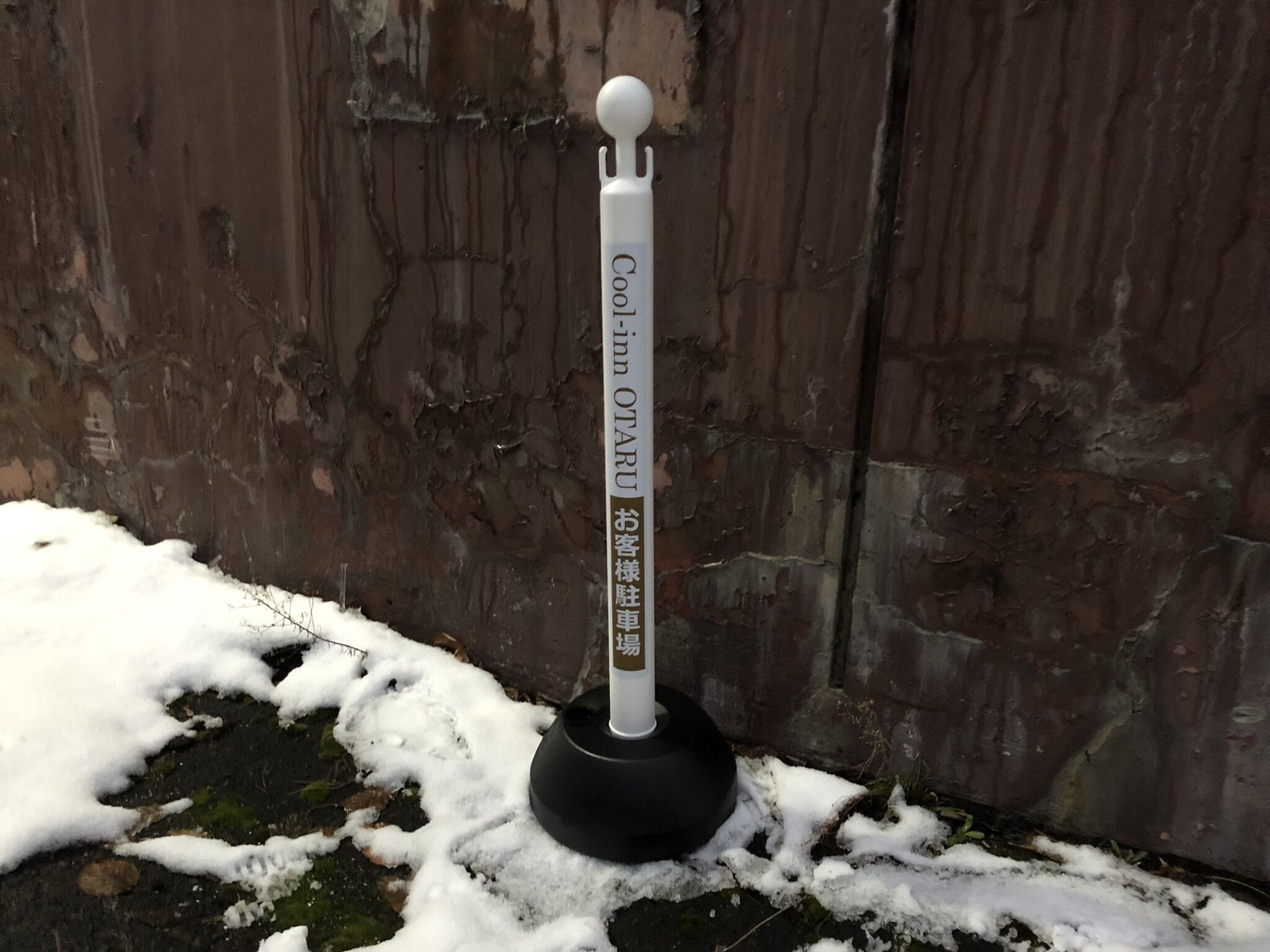 A thermometer with "Cool-in" and "OTARU" text, placed on a black base, against a wooden wall with snow on the ground.
