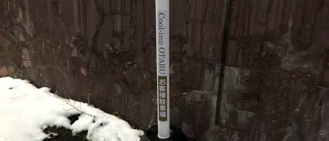 A thermometer with "Cool-in" and "OTARU" text, placed on a black base, against a wooden wall with snow on the ground.