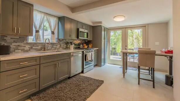 A modern kitchen with grey cabinets, a microwave, and an oven. There is a dining area with a table and chairs, and a large window providing natural light.