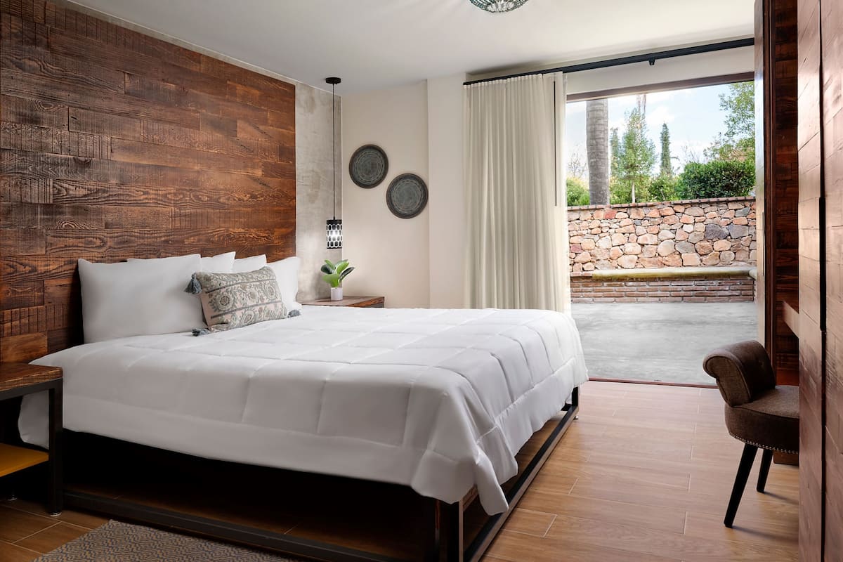 Een moderne slaapkamer met een groot bed, houten hoofdeinde en uitzicht op een stenen muur en groen buiten.