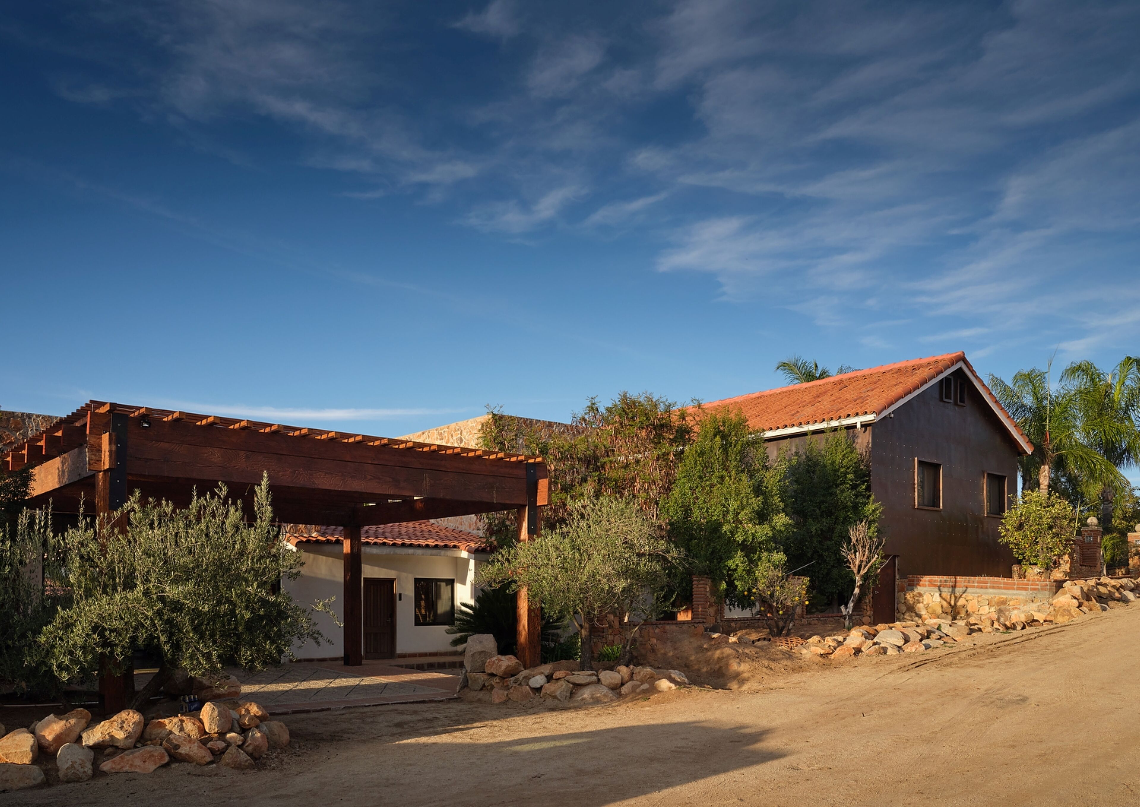 A two-story house with a red-tiled roof, a wooden pergola, and a stone wall.