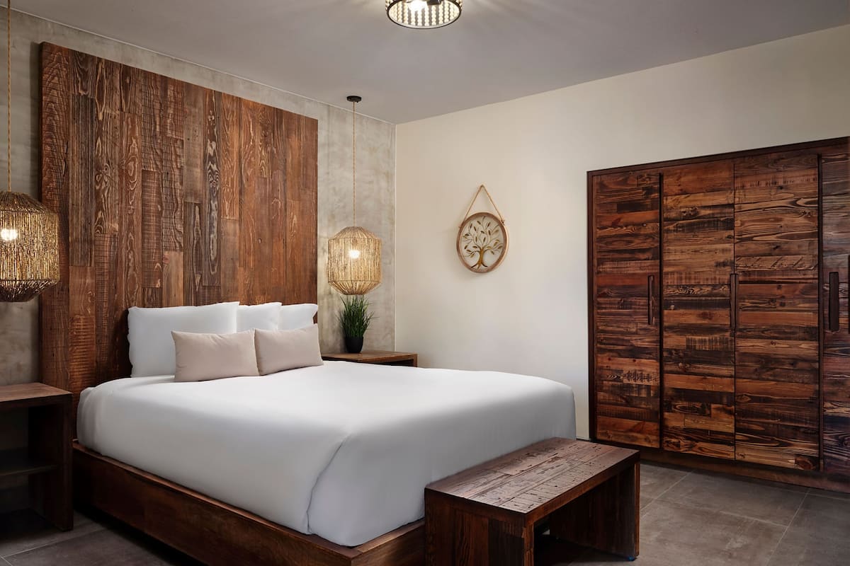 Een slaapkamer met een houten hoofdeinde, een groot bed met witte lakens, een nachtkastje, een hanglamp en een houten garderobe.