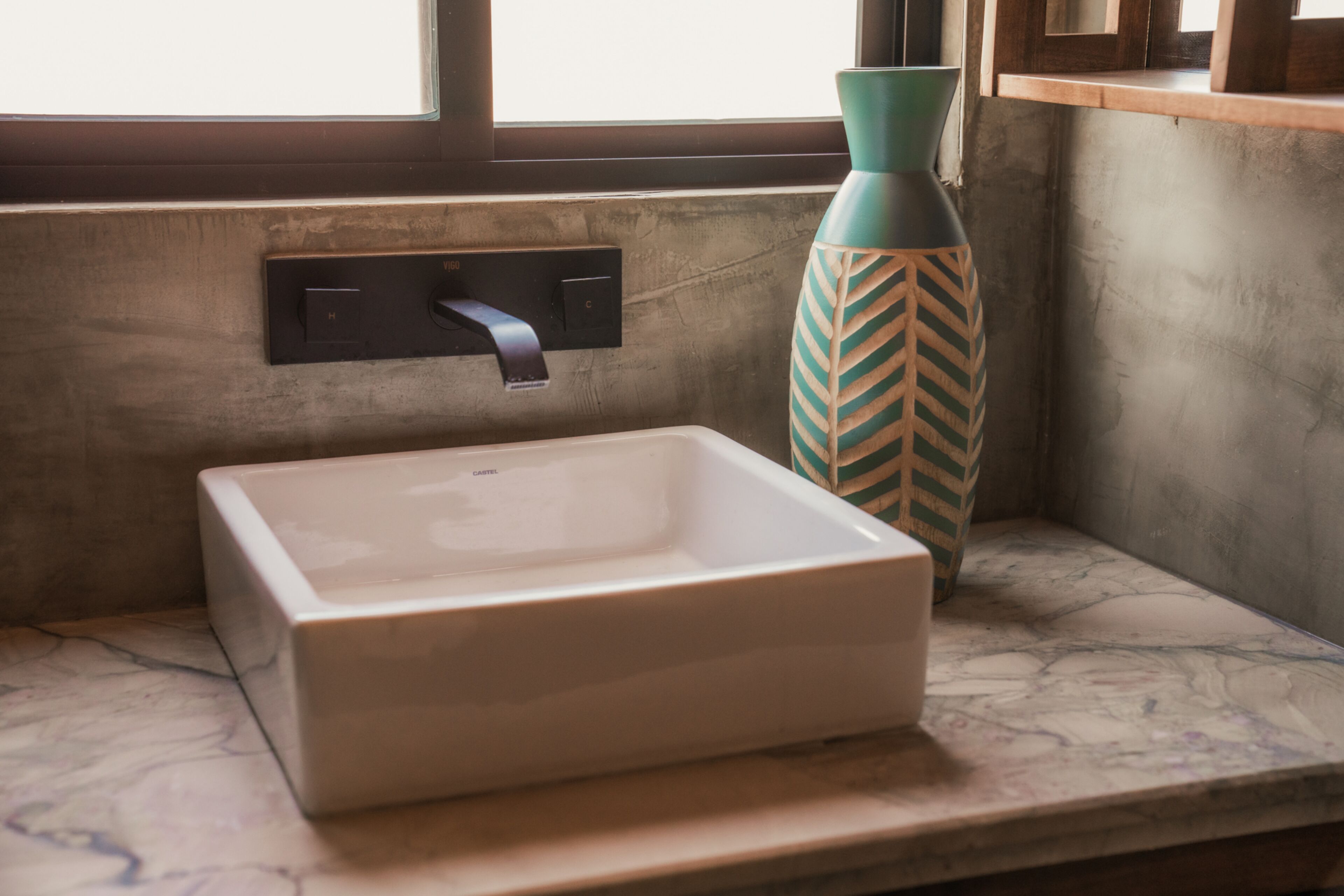 Un baño con un lavamanos blanco rectangular, un florero verde y una ventana con persianas.