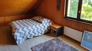 Een gezellig slaapkamer onder het dak met houten plafond, een groot bed met beddengoed met dessin, een klein nachtkastje, een radiator en een raam met uitzicht op groen.