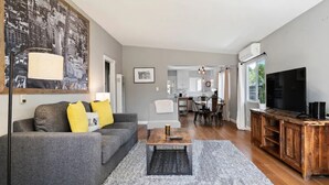 A modern living room with a grey sofa, a coffee table, a TV, and a cityscape painting on the wall. - Close to Downtown! Gorgeous MidCentury w/fire pit (Paso Robles)