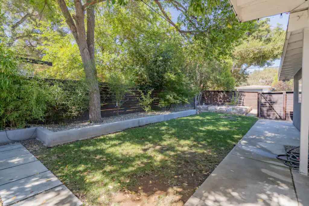 A backyard with a grassy lawn, a tree, a brick wall, and a concrete pathway.