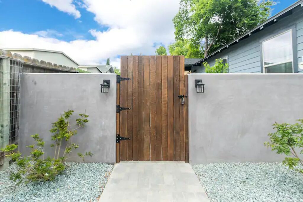 Una verja de madera con un muro de concreto, un pequeño sendero y un árbol.