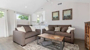 A modern living room with two sofas, a coffee table, and a rug. There are two paintings on the wall and a kitchen area visible through an open door. - Lovely Remodeled Home! Walk Downtown! w/firepit (Paso Robles)