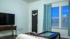 A room with a flat-screen TV on a wooden stand, a glass-top table, a bed with a striped blanket, a door with a white flower decoration, and a window with blue curtains. - Gorgeous Entire Home In Greensboro!!!! (Greensboro)