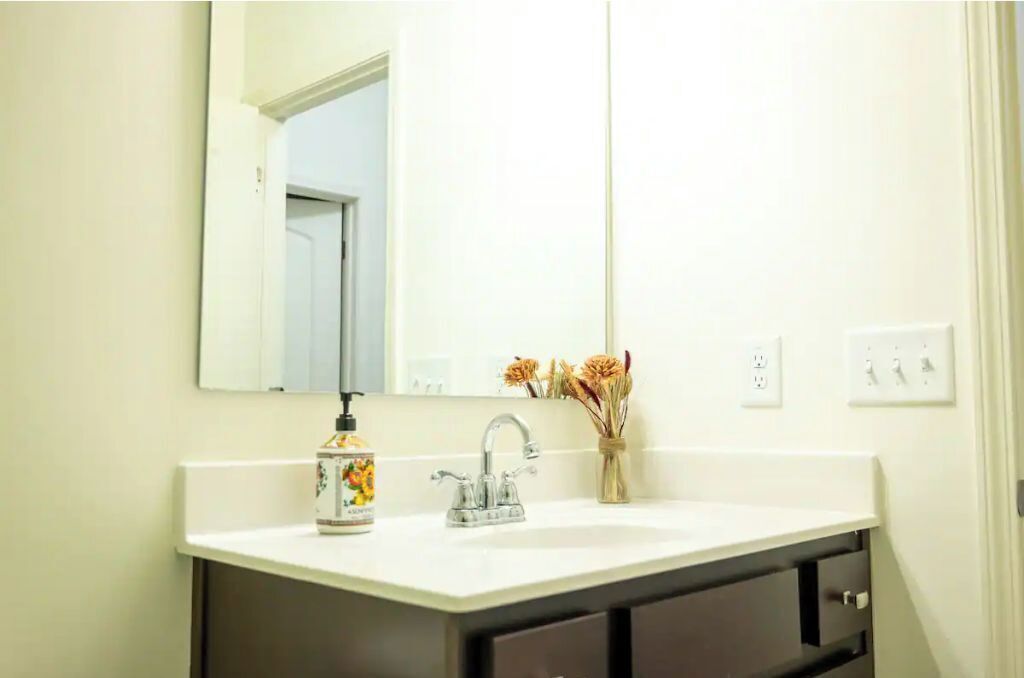 A bathroom with a white sink, a mirror, a faucet, a soap dispenser, and a vase with dried flowers.