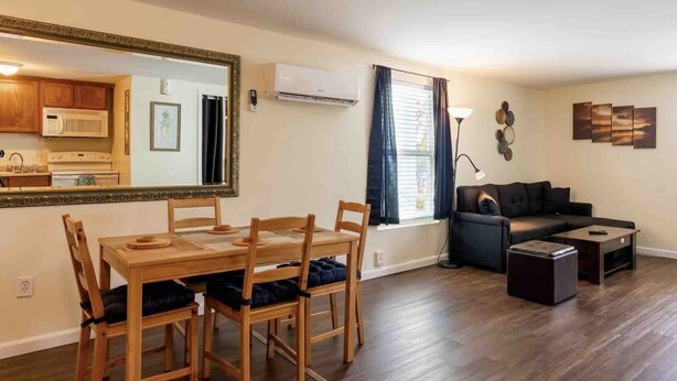 A modern living room with a dining area, wooden furniture, a large mirror, and a black sofa. - 1 BR with Ocean View - unit 1 (Fort Myers Beach)