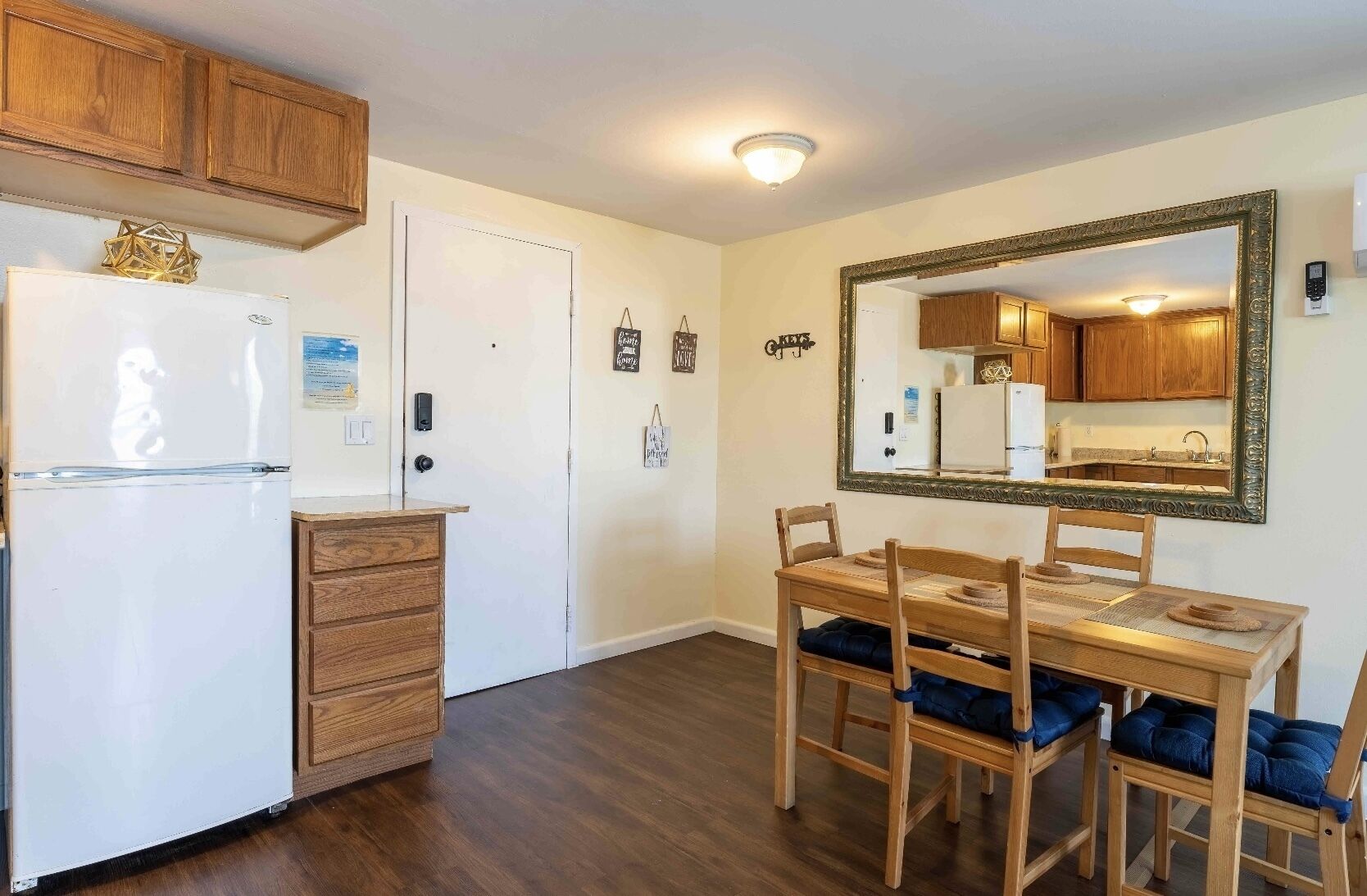 Kompakti keittiö, jossa on puiset kaapit, valkoinen jääkaappi, ruokapöytä tuoleineen ja suuri peili seinällä.