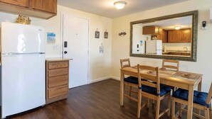 A compact kitchen with wooden cabinets, a white refrigerator, a dining table with chairs, and a large mirror on the wall. - 1 BR with Ocean View - unit 1 (Fort Myers Beach)