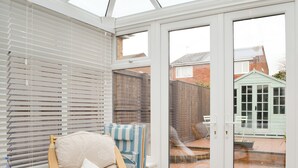 A conservatory with a wooden chair, a small table, and a view of a brick patio and a green shed. - Hollins on the Coast (Bridlington)