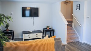 Een woonkamer met een flatscreen televisie aan de muur, een trap naar een ander niveau en een kleine tafel met een plant.