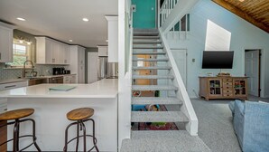 Eine geräumige Küche mit einer großen Kochinsel, ein Essbereich mit Barhocker, eine Treppe zu einer oberen Ebene sowie ein Wohnzimmer mit Fernseher und Schrank.
