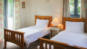 A room with two wooden beds, a nightstand with a lamp, and a window with floral curtains. - 2 bedroom accommodation in Terrington St Clements, near King&rsquo;s Lynn (Terrington St Clements, near King&rsquo;s Lynn)