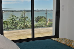 Ein Schlafzimmer mit großem Fenster, Blick auf eine Holzterrasse und ein Gewässer.