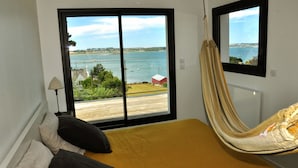 A room with a bed, a hammock, and a large window offering a view of a body of water and a red building. - "Au gré des marées", panoramic sea view, beach 300 m, town 800 m. (Carantec)