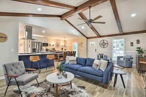 A spacious living area with a blue sofa, a grey armchair, and a coffee table. There is a kitchen in the background with a bar and a dining area. - Chic Lilburn Getaway w/Media Room + Fire Pit (Lilburn)