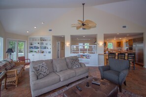A spacious living room with a large sofa, a coffee table, and a dining area with chairs. There is a kitchen visible in the background. - 13 Crooked Creek Lane (Fripp Island)