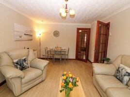 A living room with two beige sofas, a dining table with chairs, a vase of flowers, a clock on the wall, and a wooden door.