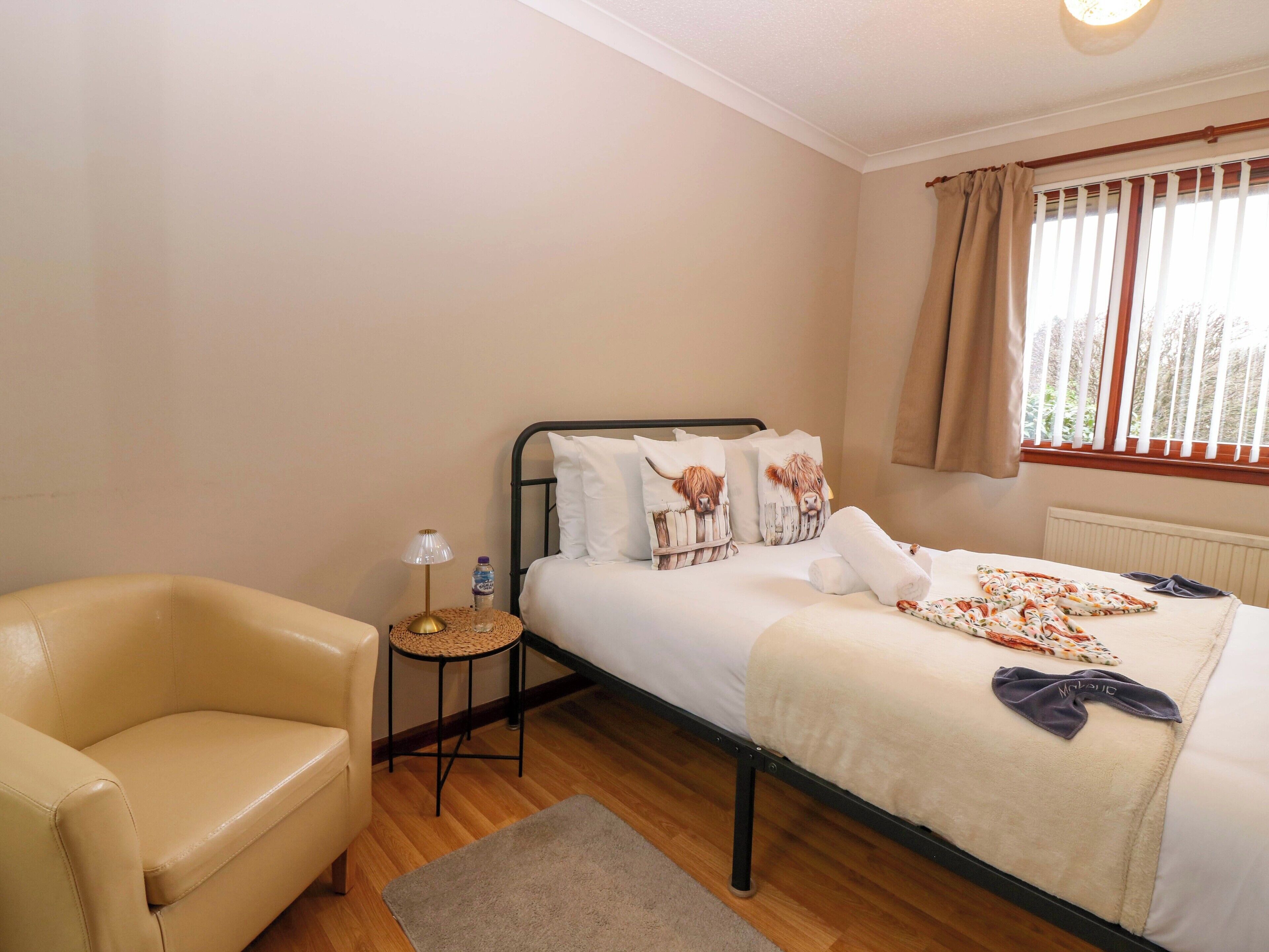A bedroom with a bed, a chair, a bedside table with a lamp, and a window with curtains.