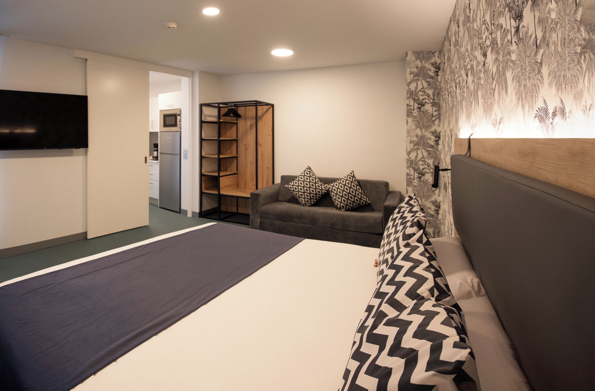 A modern hotel room with a bed, a sofa, a TV, a wardrobe, and a kitchen area.