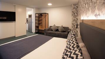 A modern hotel room with a bed, a sofa, a TV, a wardrobe, and a kitchen area.