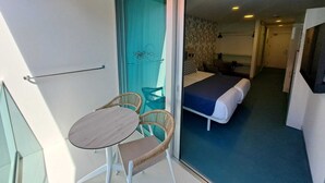Un balcón con mesa y sillas, y un ventanal corredera que da a un dormitorio con cama y sillón.