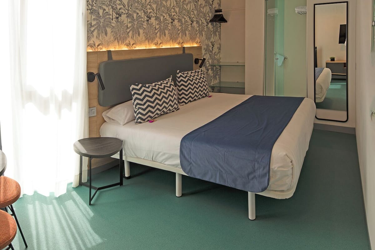 A hotel room with a bed, a small table, a chair, a mirror, and a door leading to another room.