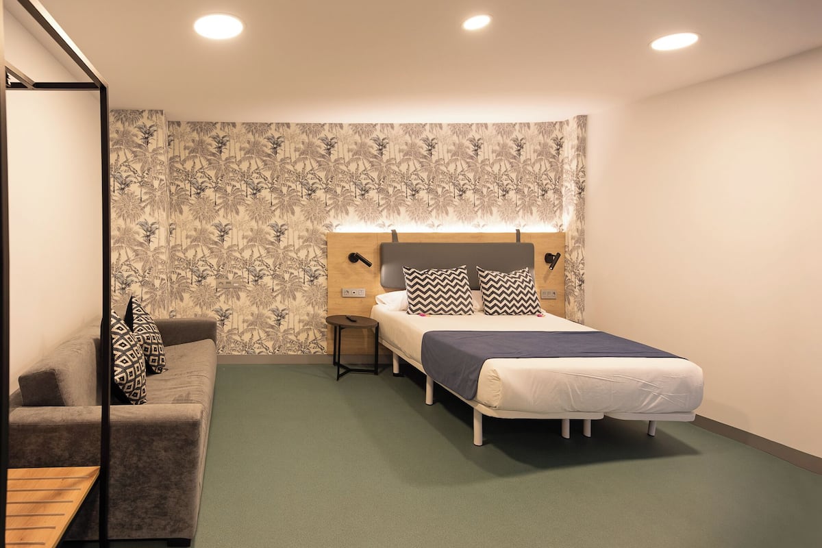 A bedroom with a bed, a nightstand, a sofa, and a wall with a tropical wallpaper.