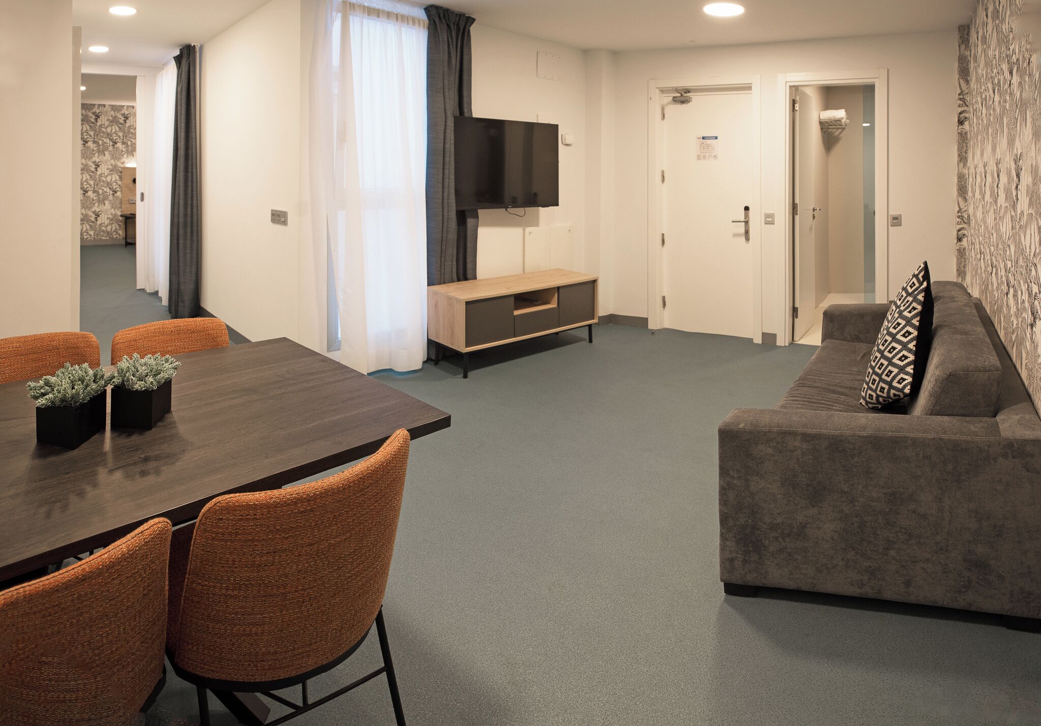 A modern living room with a dark wooden dining table, orange chairs, a grey sofa, a flat-screen TV, and a small wooden cabinet.