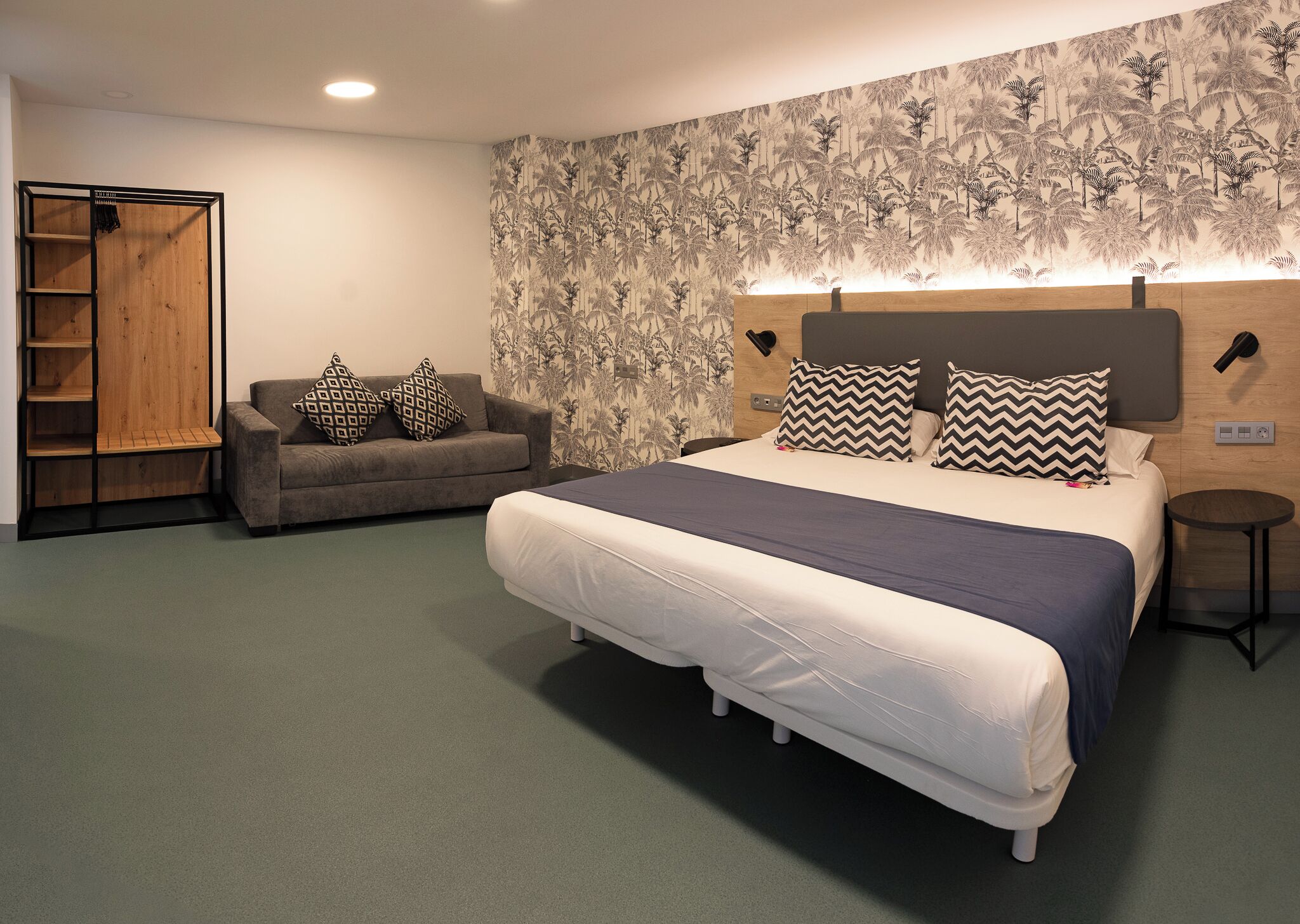 A bedroom with a bed, a sofa, a wardrobe, and a wall with a tropical wallpaper.