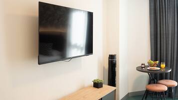 Una habitación de hotel moderna con un televisor de pantalla plana montado en la pared, una mesita de consola de madera, una pequeña planta en maceta, un calentador pequeño, una mesa redonda con dos sillas y una cortina oscura.