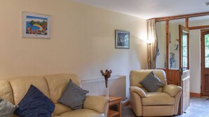 Un salon avec un canapé beige, un fauteuil plus petit et une table basse en bois. Deux tableaux encadrés sont accrochés au mur, un radiateur se trouve sous le canapé et une fenêtre avec des rideaux est située à droite.